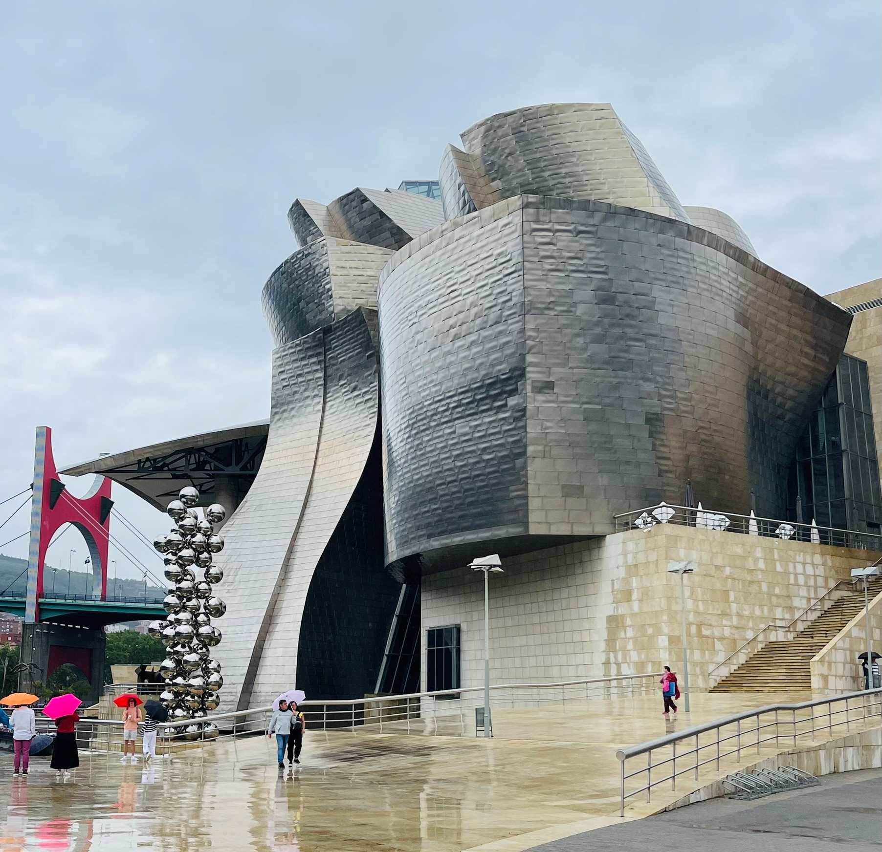 Silver/gray modern building with bridge in back with people with umbrellas in front in Bilbao, Northern Spain